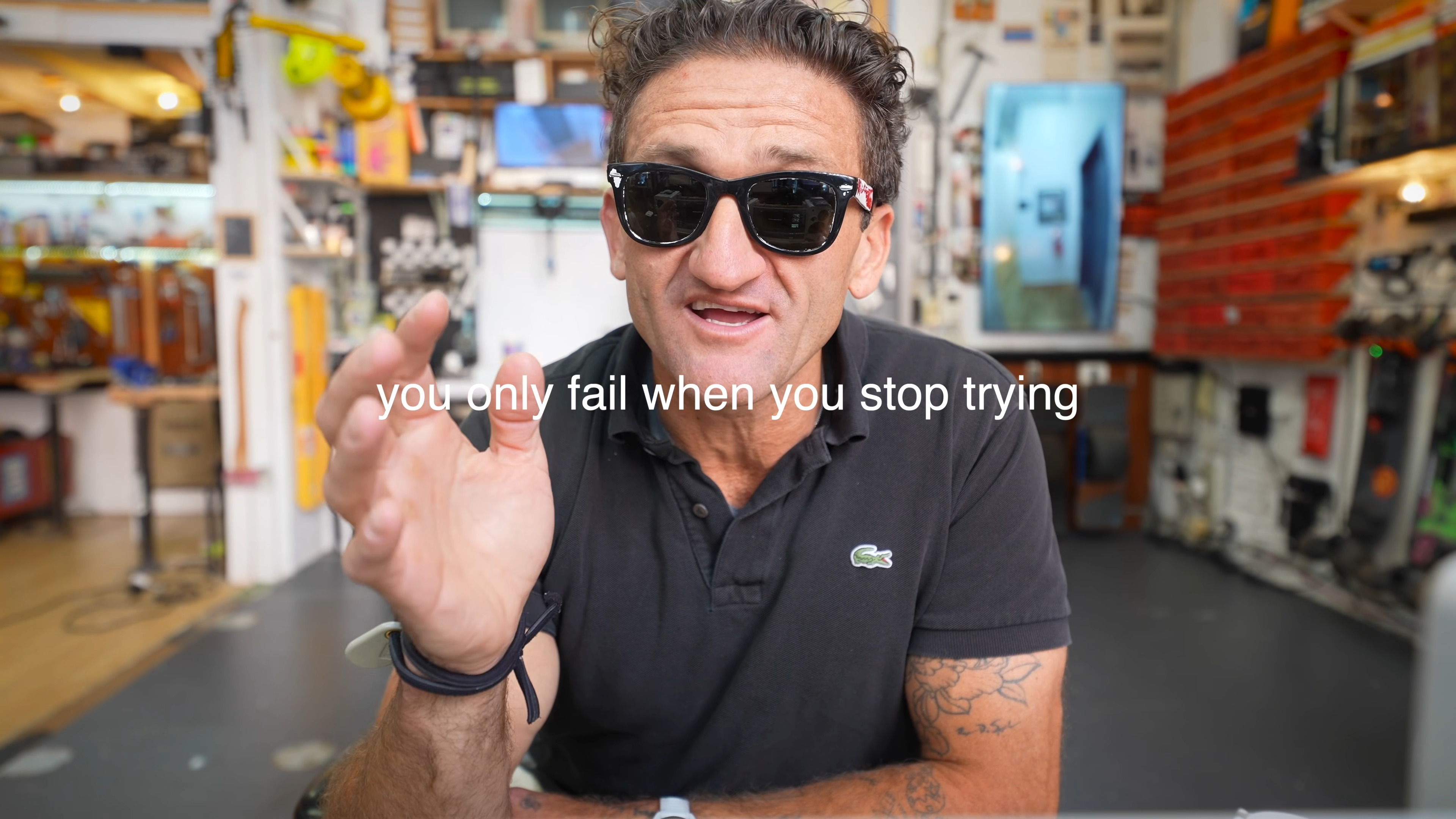 A man wearing black sunglasses and a black polo shirt with a Lacoste logo is speaking to the camera in what appears to be a workshop or studio setting. He has his right hand raised as if gesturing while talking, and there is a visible tattoo on his left forearm. The background is filled with various objects, equipment, and shelves, indicating a creative or workshop environment. The text "you only fail when you stop trying" is superimposed over the image.
