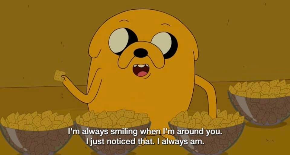 An animated scene from the show Adventure Time featuring the character Jake the Dog. Jake is a yellow, anthropomorphic dog with a large, round head and small black eyes. He has a wide, happy expression with an open mouth, showing his tongue. He is holding a small cracker or snack in his right hand. In front of him are three bowls filled with small, square crackers. The background is a solid, warm brownish-orange color.