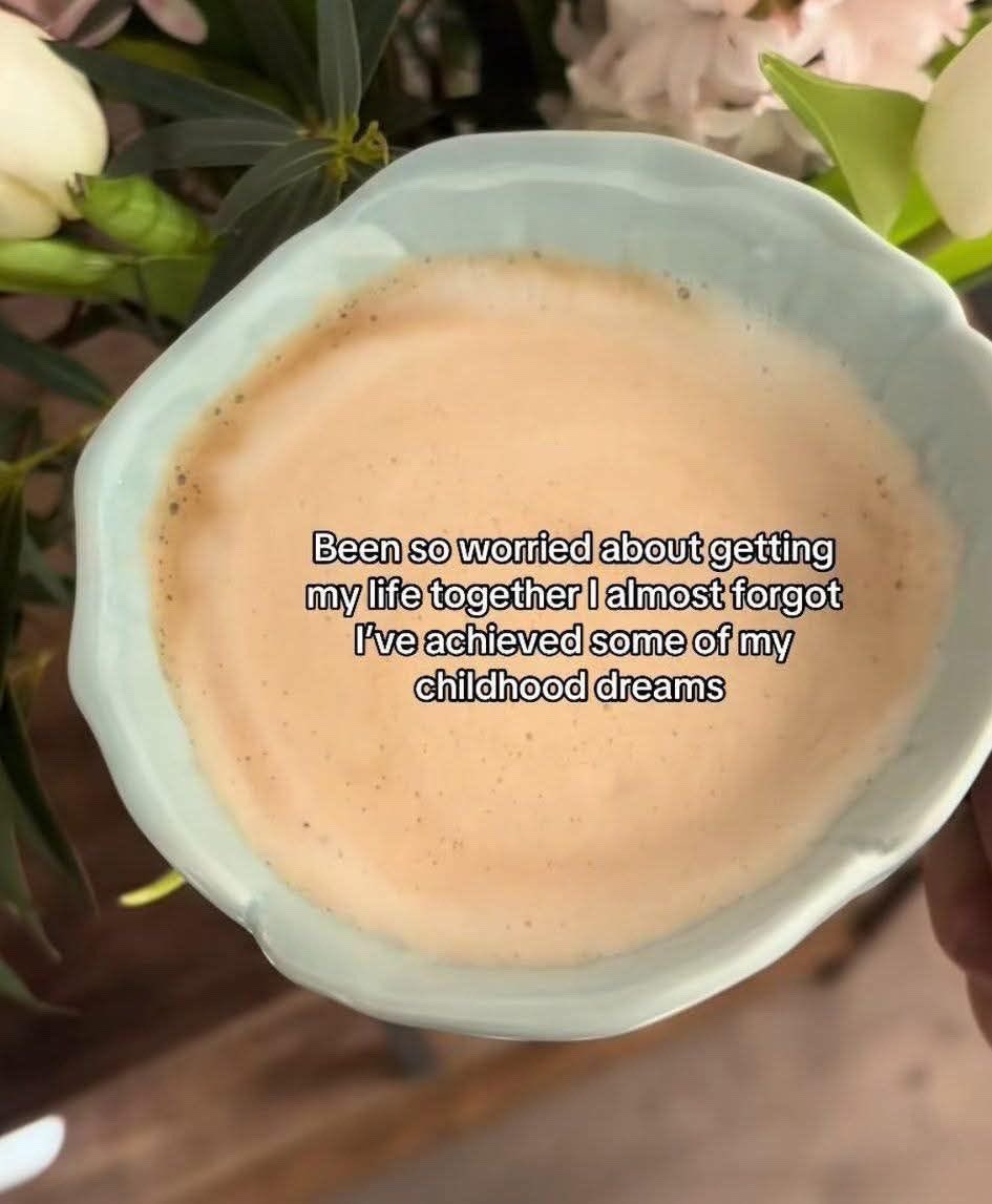 A top-down close-up shot of a light blue, wavy-edged ceramic bowl filled with a creamy, tan-colored liquid, possibly a latte or smoothie. In the background, there are soft-focus white and pink flowers with green leaves. Centered over the liquid is a text overlay with a sentimental message.