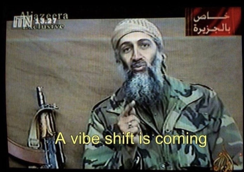 A low-resolution, meme-style image featuring a man with a long grey beard and a skullcap, wearing camouflage clothing. He is gesturing with one finger raised. In the background, a rifle is visible to the left. The image appears to be a screen capture from an Al Jazeera news broadcast, indicated by the logos in the top corners. Large yellow text is overlaid at the bottom of the image.