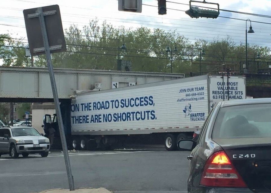 A street-level shot showing a white semi-truck trailer driving under a bridge. The trailer has blue text that reads 'ON THE ROAD TO SUCCESS, THERE ARE NO SHORTCUTS.' and branding for 'SHAFFER TRUCKING'. In the background, there are green trees, utility lines, and a street light. In the foreground, the rear of a black car is visible on the right, and a white SUV is driving on the left. A grey street sign post is partially visible in the left foreground.