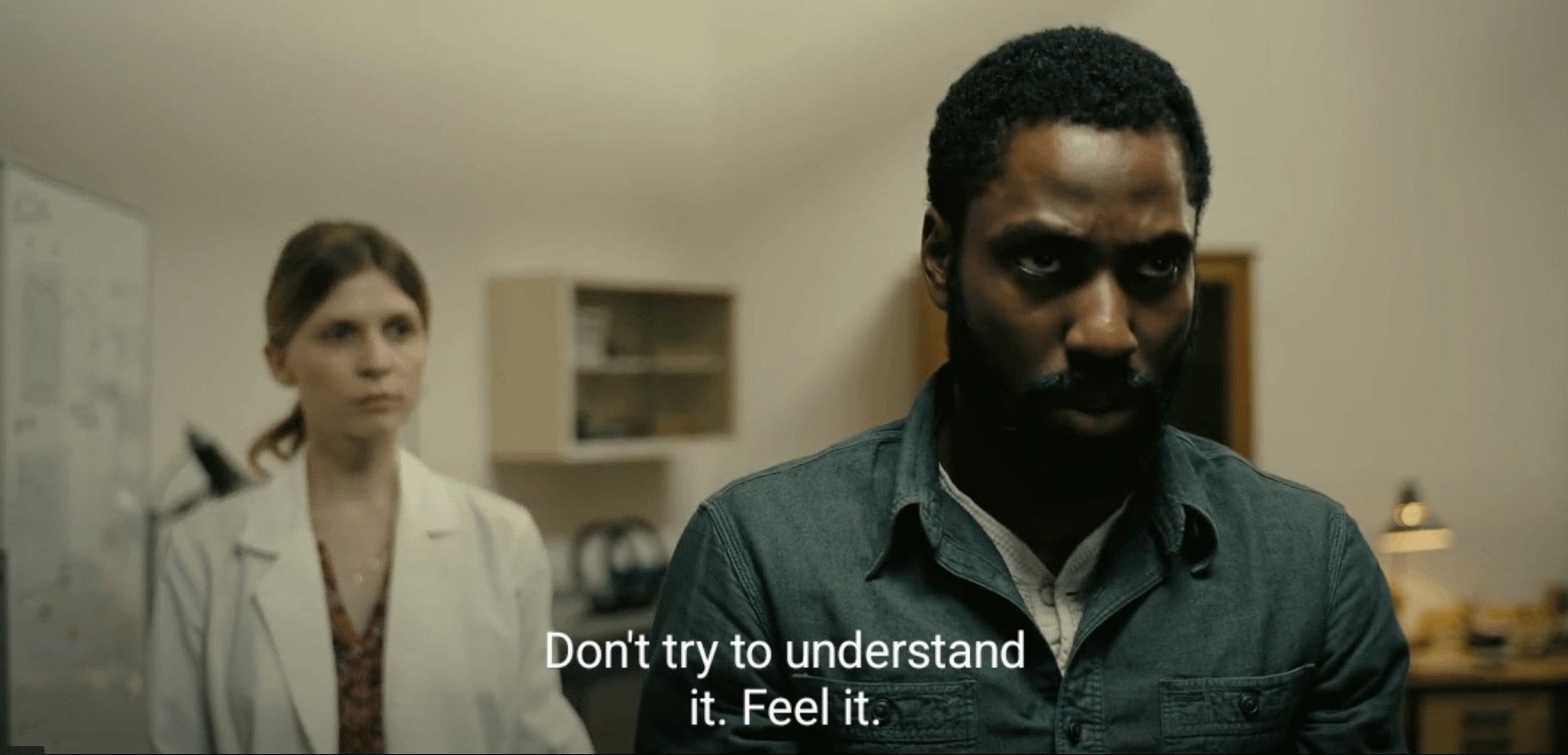 A cinematic medium shot featuring two people in an indoor setting. In the foreground, a Black man with a beard and a serious, intense expression is wearing a dark denim shirt over a white undershirt. In the blurred background, a white woman with light brown hair, wearing a white lab coat over a patterned top, looks on with a neutral expression. The room appears to be an office or a laboratory with a wooden cabinet and a lamp visible. Subtitles at the bottom of the frame read 'Don't try to understand it. Feel it.'
