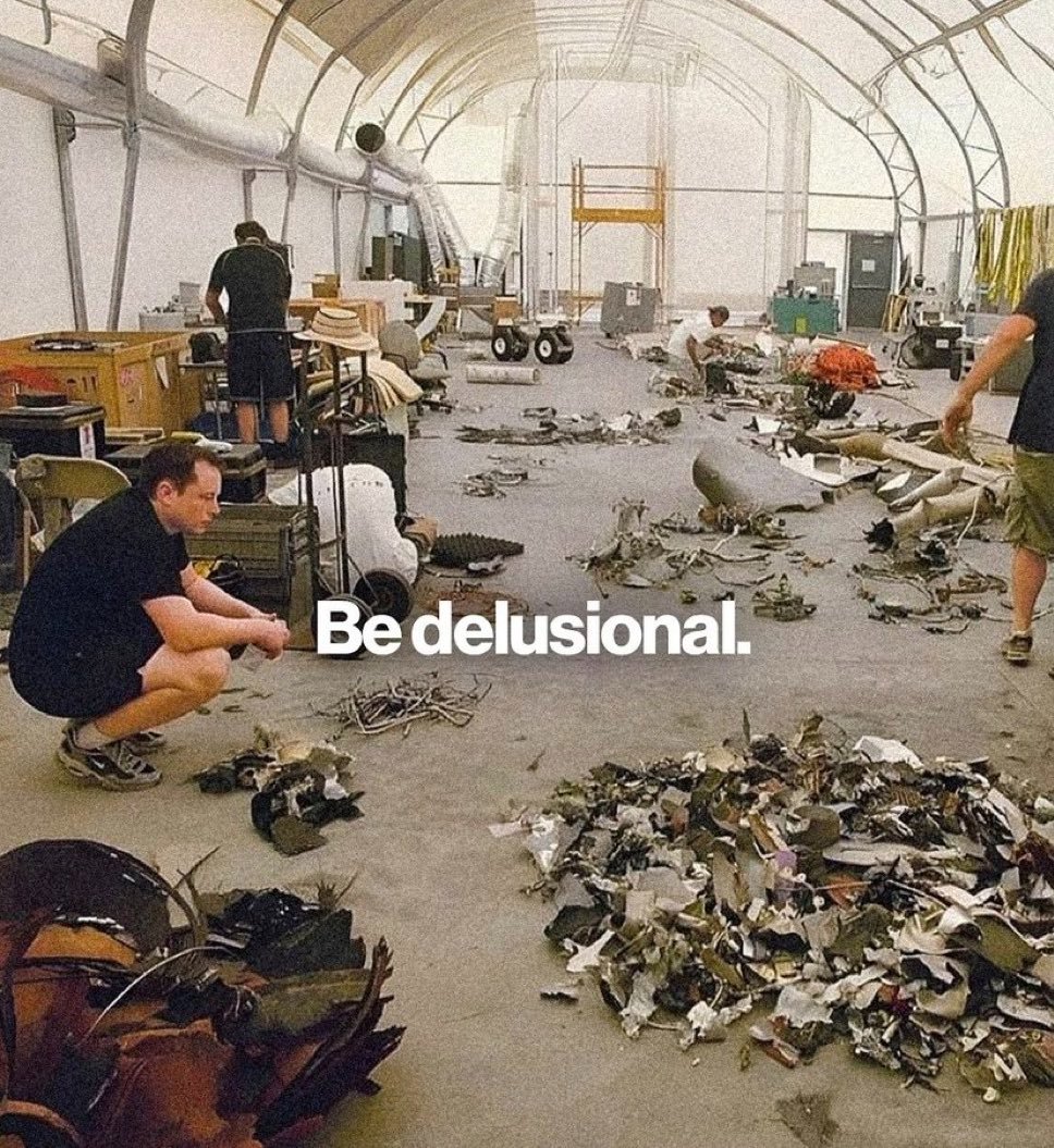 A photograph of a large, hangar-like industrial space filled with scattered metal debris and wreckage on the floor. Several people are seen moving around the wreckage, appearing to be inspecting or cleaning up. In the foreground, a man in a black shirt is crouching down, looking at the debris. The image has white text overlaid in the center that reads 'Be delusional.'