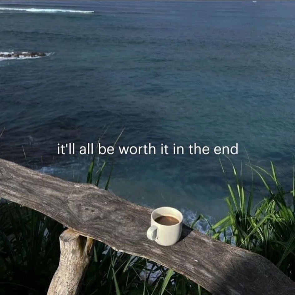 A serene coastal scene featuring a large piece of weathered wood acting as a ledge or table overlooking a vast, blue ocean. A single white mug filled with coffee sits on the wooden ledge. Green coastal plants are visible in the foreground and around the wood. The text 'it'll all be worth it in the end' is centered across the middle of the image.