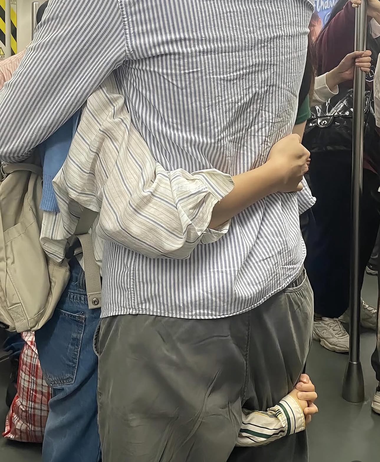 A close-up shot of people standing in a crowded public space, likely a subway or train. In the center, a person wearing a blue and white vertical striped shirt and grey trousers is being groped by two different hands: one hand is grabbing their waist from the side, and another hand is reaching underneath the back of their trousers. Other commuters, including someone with a backpack and someone wearing a dark jacket and white sneakers, are visible in the periphery.