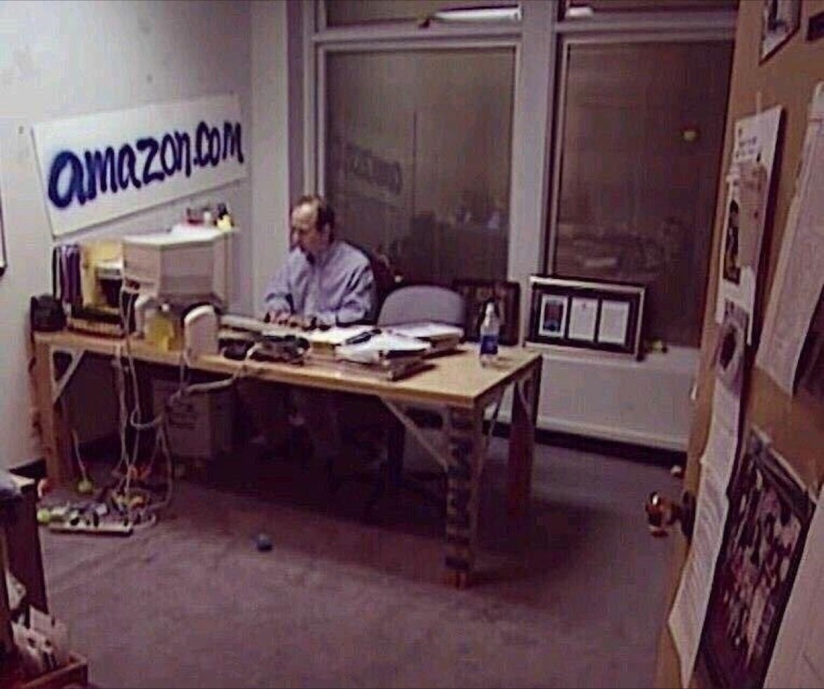 A low-resolution, somewhat grainy photograph of a man sitting at a desk in an office setting. The man, who appears to be Jeff Bezos, is wearing a light-colored button-down shirt and is working at a desk cluttered with various items including a computer, papers, and a water bottle. On the wall behind him, there is a large sign that reads 'amazon.com'. To the right of the man, there is a window looking out into another area, and several posters or papers are pinned to the wall on the far right. The office appears to be a modest, early-stage workspace.