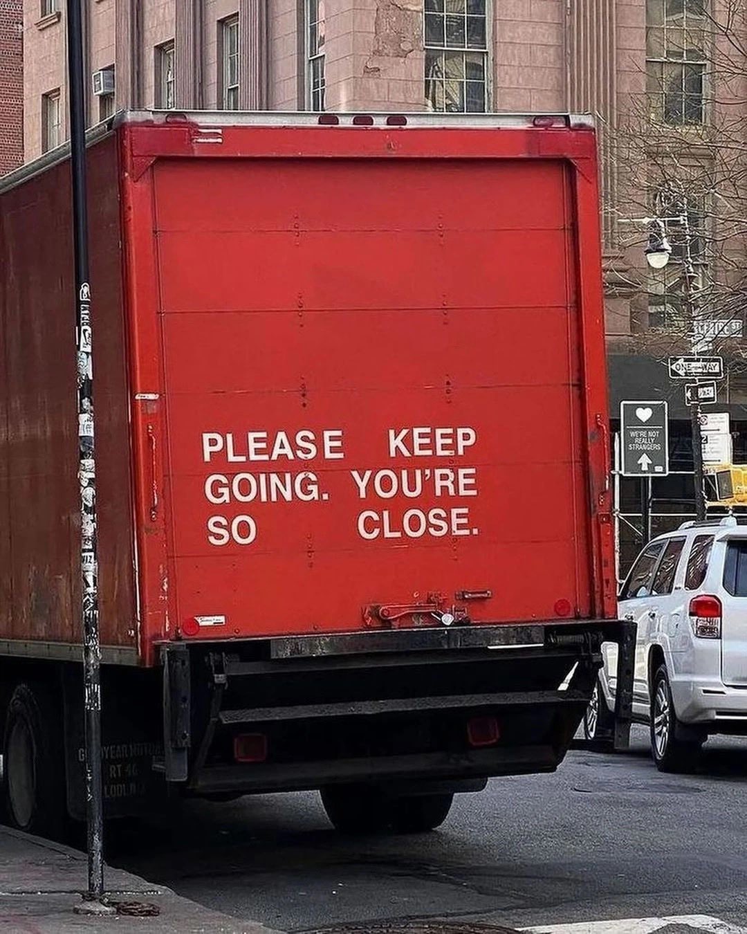 A vertical, medium shot shows the rear of a large red box truck parked on a city street. The truck's back door features large, white, sans-serif text that reads, 'PLEASE KEEP GOING. YOU'RE SO CLOSE.' To the left of the truck is a black pole with various stickers. To the right, a white SUV is partially visible, followed by a street scene with a building, street signs (including a 'ONE WAY' sign), and a small sign with a heart that says 'WE'RE NOT REALLY STRANGERS'. The background shows a multi-story brick building under a clear sky.