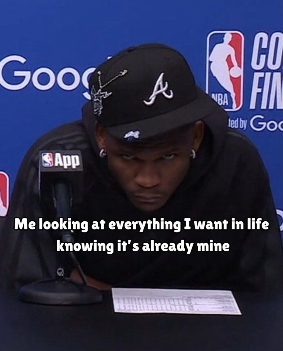 A meme featuring a close-up photo of NBA player Trae Young wearing a black hoodie and a black Atlanta Hawks hat. He has a serious, intense, or focused facial expression, staring directly at the camera. He is seated in front of an NBA backdrop with various logos, including Google and the NBA logo. Superimposed white text on the image reads: 'Me looking at everything I want in life knowing it's already mine'.