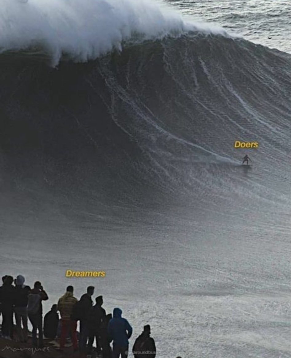 A vertical meme image featuring a massive, towering ocean wave. In the foreground, a group of people stands on a shore or ledge, labeled with the text 'Dreamers'. Further out, a lone surfer is riding the face of the giant wave, labeled with the text 'Doers'. The image serves as a metaphor for taking action versus just thinking about it.