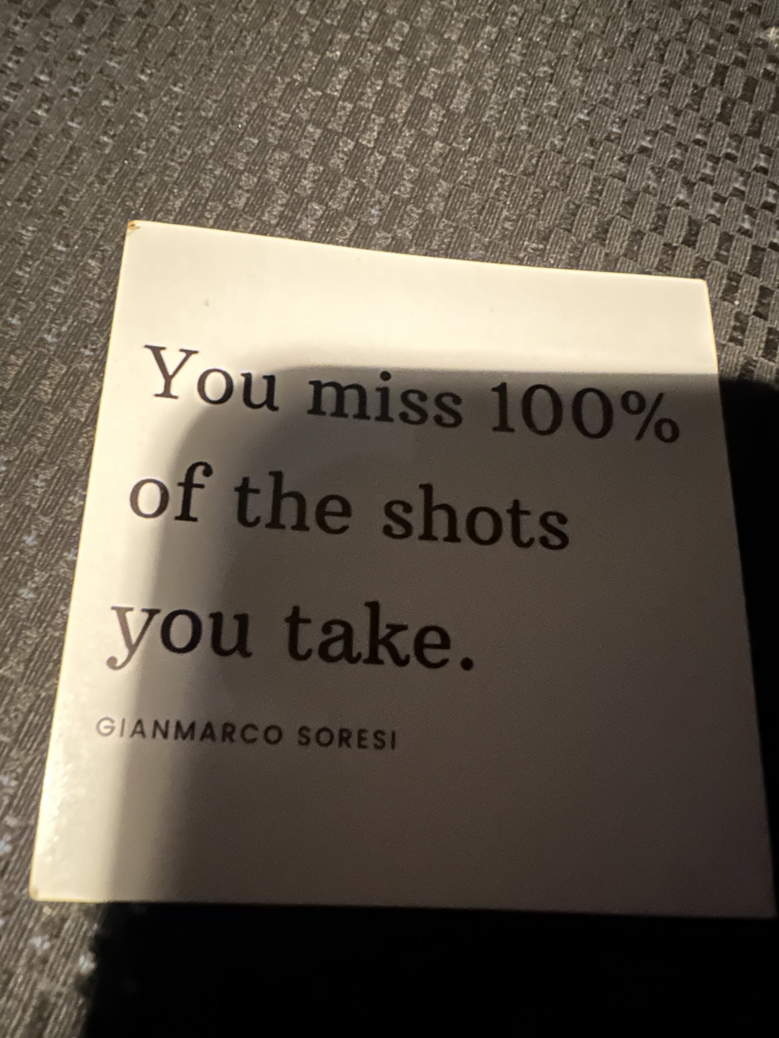 A close-up, slightly angled shot of a white square card resting on a dark, textured grey surface. The card features a quote printed in black serif typography. The lighting creates a diagonal shadow across the card.