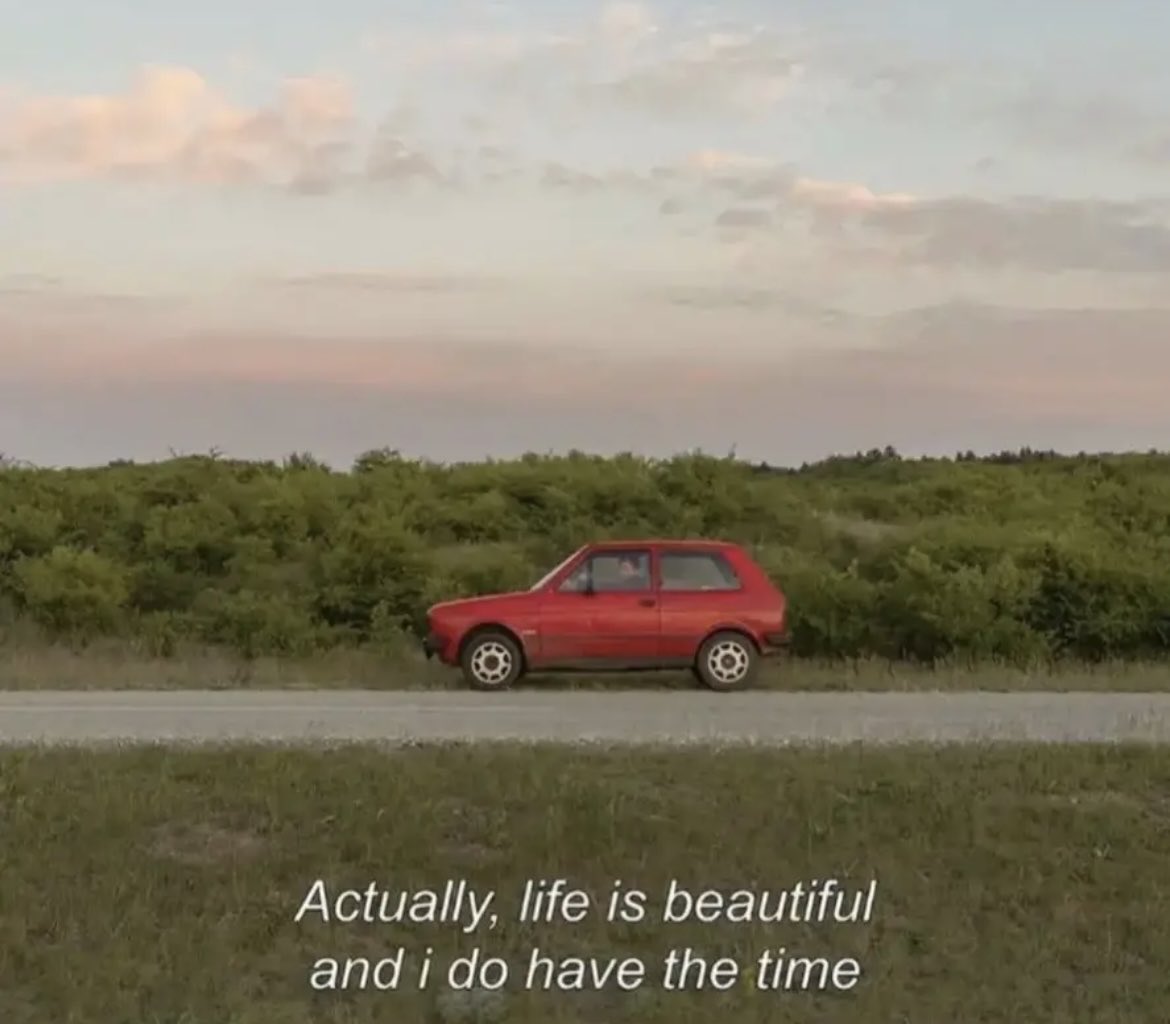 A landscape photo at dusk or dawn featuring a small red car parked on the side of a road. Behind the car is a dense line of green bushes and trees. The sky is filled with soft, pastel-colored clouds in shades of pink and grey. In the foreground, there is a grassy field. White text is overlaid at the bottom of the image.