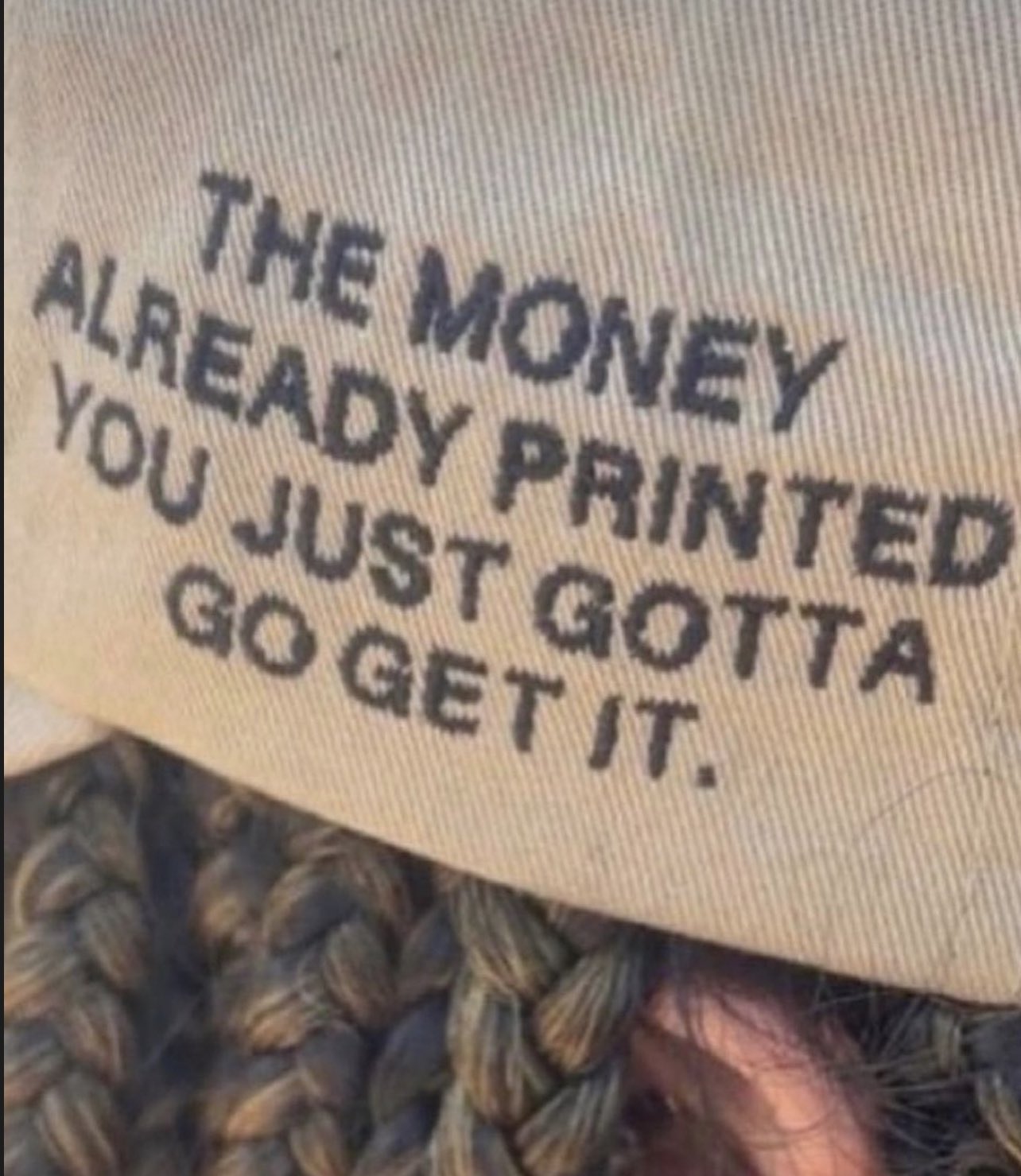 A close-up shot of a tan or beige fabric, likely part of a hat, featuring printed text in black capital letters. The text is slightly angled. Below the fabric, some braided hair or a textured material is visible.