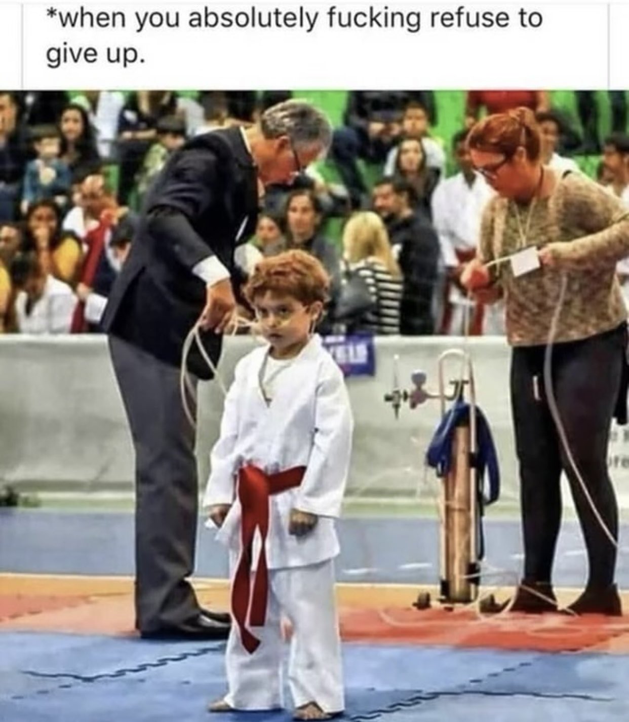 A meme featuring a photo of a young child in a white karate gi with a red belt. The child has a small cut or blood on their nose and an intense, determined facial expression. In the background, two adults are tending to the child; a man in a suit is holding what appears to be medical tubing, and a woman is standing near an oxygen tank. The setting is a martial arts competition mat with a crowd of spectators in the background. The meme text at the top reads: '*when you absolutely fucking refuse to give up.'