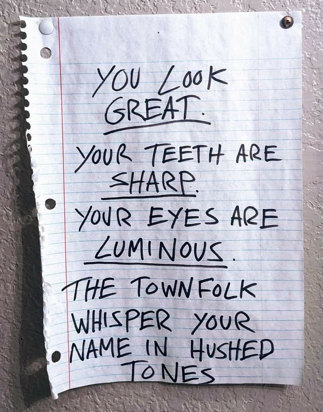 A vertical shot of a piece of white lined notebook paper taped to a textured, light-colored wall. The paper has handwritten text in black marker. The text is written in all caps and conveys a slightly eerie or unsettling tone through its compliments. The paper has perforated edges on the left side and a red margin line.
