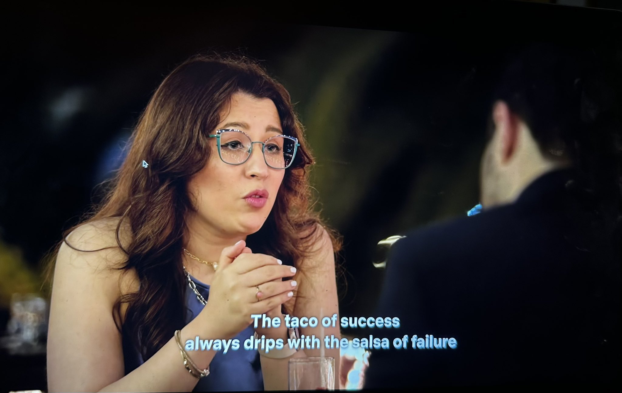 A close-up shot of a woman with long brown hair and glasses, wearing a blue top and jewelry. She is gesturing with her hands while speaking to a man, whose back is partially visible in the foreground. Subtitles at the bottom of the frame read, 'The taco of success always drips with the salsa of failure.' The image appears to be a photograph of a screen displaying a video.