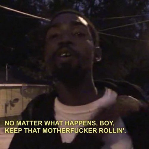 A low-quality, dimly lit still image of a Black man looking towards the camera at night. He has a beard and is wearing a dark jacket over a white shirt. At the bottom of the image, there is white text overlaid on the frame.