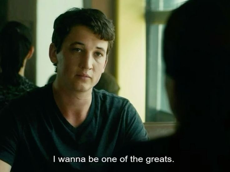 A cinematic medium shot of actor Miles Teller looking towards another person (seen from the back/silhouette) in what appears to be an indoor setting. He has a serious, contemplative expression and is wearing a dark V-neck t-shirt. The background is softly blurred, showing a hint of a window and another person's silhouette.
