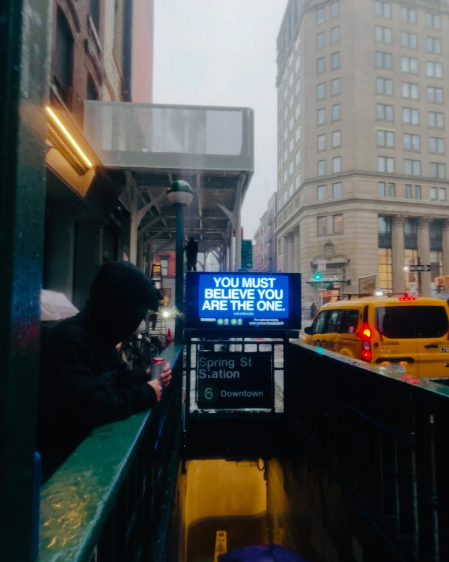 A vertical, slightly low-angle shot taken on a rainy or overcast day in a city. In the foreground, a person wearing a black hoodie is partially visible, leaning against a railing next to a subway entrance. They appear to be holding a small can. The subway entrance features a digital sign displaying the text 'YOU MUST BELIEVE YOU ARE THE ONE.' below it, a sign reads 'Spring St Station 6 Downtown'. A yellow taxi is driving past on the street to the right. The background consists of tall city buildings and an elevated walkway structure under a gray, misty sky.