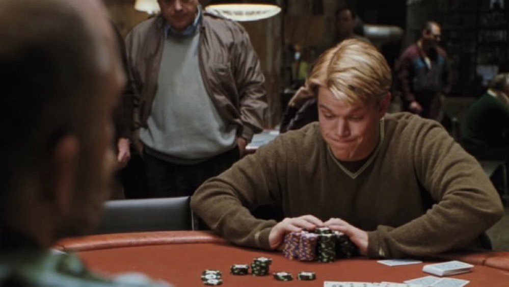 A medium shot from a film showing a blond man in a brown sweater sitting at a poker table, looking down with a focused or slightly tense expression. In front of him are several stacks of multicolored poker chips. In the foreground, the blurred back of another person's head is visible, suggesting a first-person perspective from across the table. The background is dimly lit, showing other people in a casino or card room setting, including a man in a brown jacket standing behind the player.