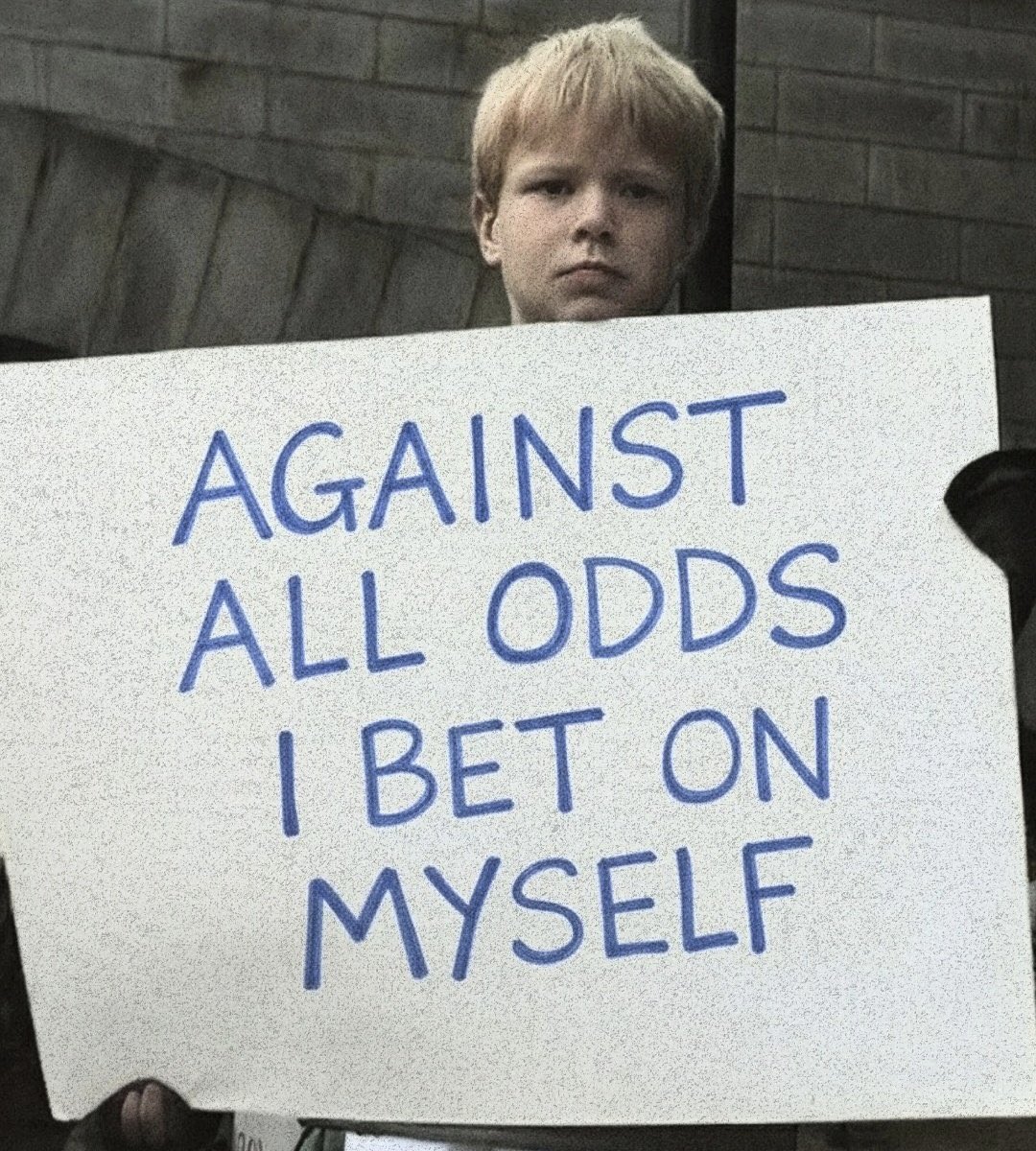 A close-up shot of a young boy with blond hair holding a large white sign. The boy has a serious facial expression and is looking directly at the camera. The sign is written in blue capital letters and reads 'AGAINST ALL ODDS I BET ON MYSELF'. The background consists of a brick wall and some stairs.