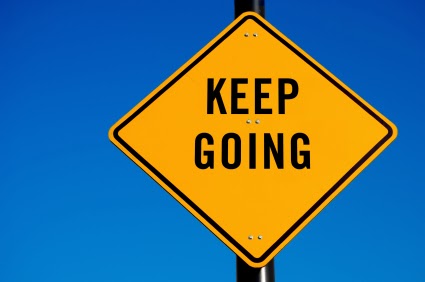 A yellow diamond-shaped sign mounted on a black pole against a solid blue background. The sign contains the text 'KEEP GOING' in bold, black, capital letters.