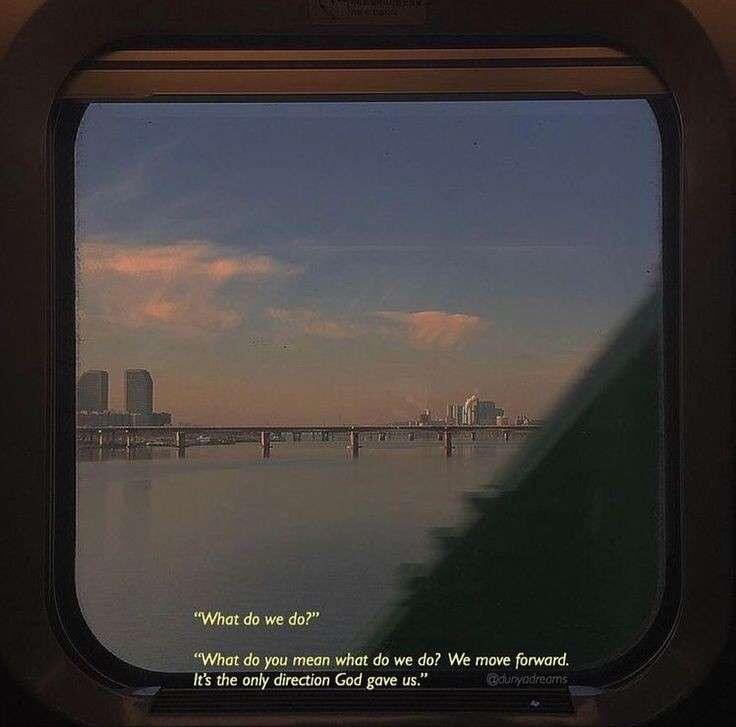 A view through a rounded rectangular window, likely from a train or vehicle, looking out over a calm body of water toward a distant city skyline at dusk or dawn. The sky features soft orange and blue hues with wispy clouds. A long bridge stretches across the water in the distance. A dark, blurry shape, possibly a part of the vehicle or a tree, obscures the bottom right corner of the view. Text is overlaid at the bottom of the image.