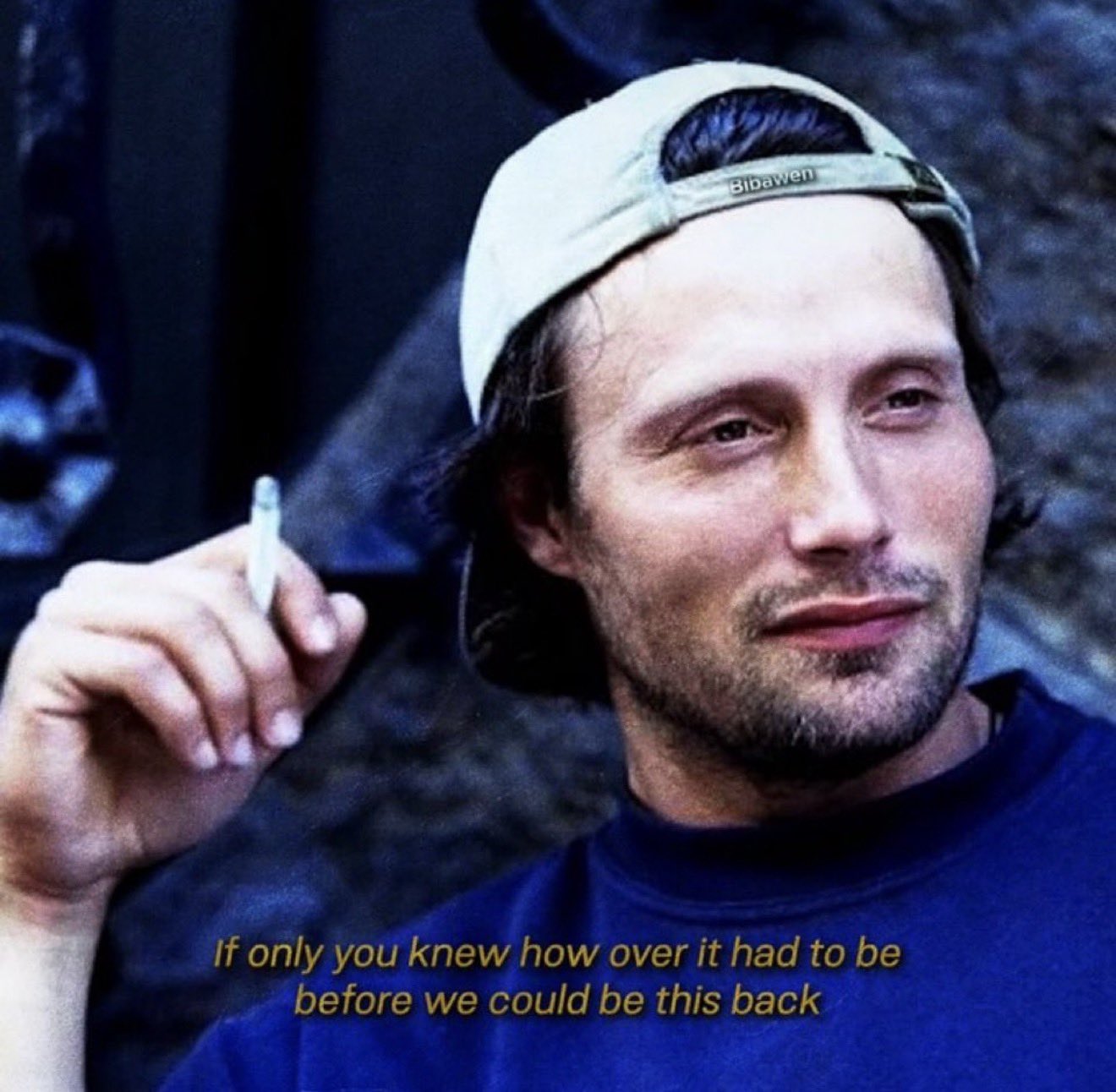 A close-up portrait of a man with dark hair and light stubble, wearing a light-colored baseball cap backwards and a navy blue t-shirt. He is looking slightly to the side with a subtle, perhaps wistful, expression. In his right hand, he holds a lit cigarette. The background is dark and out of focus. Yellow italicized text is overlaid at the bottom of the image.