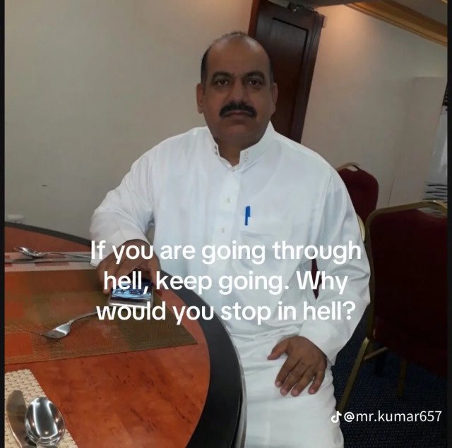 A medium shot of a middle-aged man with dark hair and a mustache, wearing a white button-down shirt. He is sitting at a wooden dining table in what appears to be a dining room or restaurant. The background shows a door and a chair. Superimposed on the image is a motivational quote in white text.
