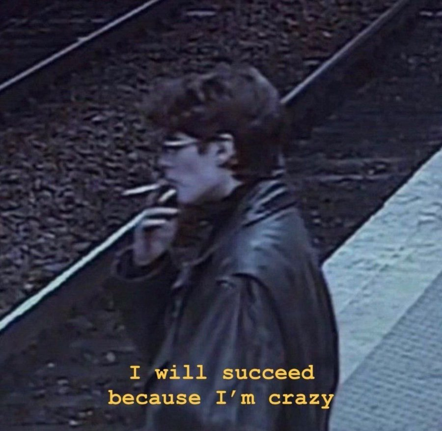 A low-resolution, grainy, dark-toned photograph of a person with dark hair and glasses, wearing a black leather jacket. The person is shown in profile, smoking a cigarette. The background appears to be a textured, dark outdoor surface like gravel or asphalt next to a light-colored pavement. Yellow text is overlaid at the bottom of the image.