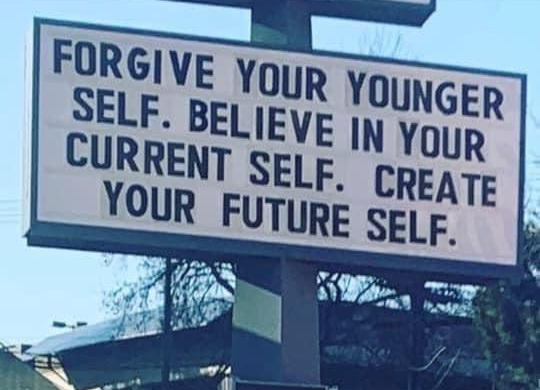 A rectangular sign mounted on a pole against a clear blue sky. The sign features large, bold, black capital letters on a white background with a motivational message.
