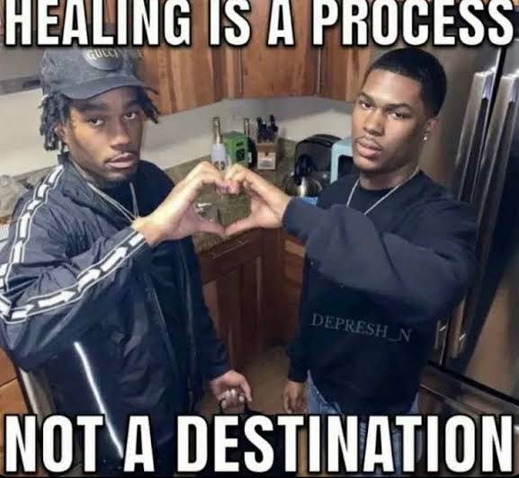A meme featuring two young men. One man on the left, wearing a dark jacket and a grey baseball cap, is making a heart shape with his hands. The other man on the right, wearing a dark sweatshirt, is also participating in making the heart shape with his hands. The background appears to be an indoor setting, possibly a kitchen. Large white text is overlaid on the image, reading 'HEALING IS A PROCESS' at the top and 'NOT A DESTINATION' at the bottom.