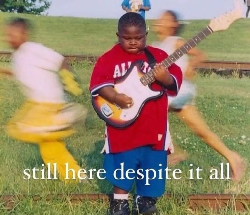 A low-resolution, slightly blurry photograph featuring a young Black boy standing centrally in the foreground. He is wearing a red jersey with white lettering and blue shorts, and he is holding and appearing to play an electric guitar. The boy has a serious expression. In the background, two other children are captured in motion blur, appearing to run past him. The setting is an outdoor grassy area near some railroad tracks. Overlaid at the bottom of the image is the text 'still here despite it all' in a white, serif font.