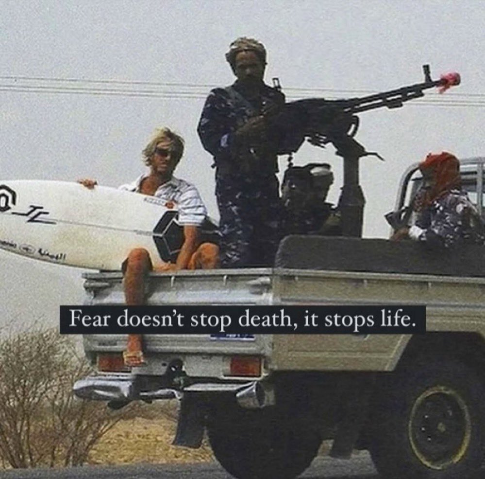 A meme featuring a low-quality, grainy photograph of three men in the back of a white pickup truck. One man is sitting on the edge holding a white surfboard; a second man stands in the middle holding a large machine gun; and a third man is partially visible in the front passenger seat wearing a red head covering. The setting appears to be a dry, desert-like environment. A black text box is overlaid on the bottom half of the image with white text.