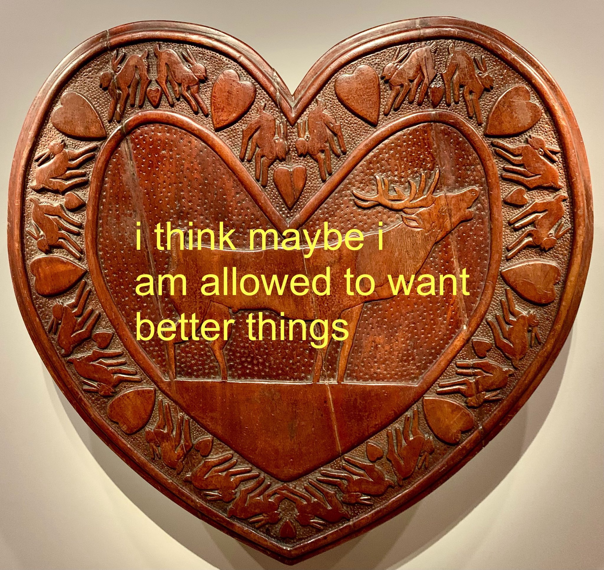 A close-up photograph of a heart-shaped wooden carving. The carving features a large deer in the center and is bordered by smaller carvings of rabbits and hearts. Overlaid on the center of the image in bright yellow sans-serif font is the text: 'i think maybe i am allowed to want better things'.