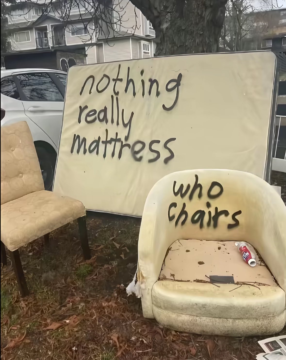 A collection of discarded furniture sits on a patch of grass and dead leaves outdoors. In the background, there is a white car and residential houses. The furniture includes a large, upright mattress leaning against a tree, a tan upholstered chair on the left, and a worn-out, white armchair in the foreground. The mattress and the armchair both have humorous, spray-painted text on them.