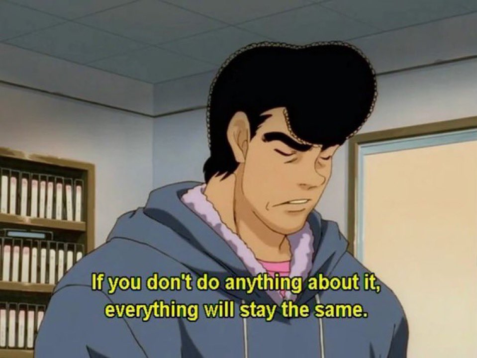 An anime-style still of a man with large, black, pompadour-style hair and thick eyebrows. He is wearing a blue hooded sweatshirt with a light purple fuzzy lining. He has a serious or contemplative expression on his face. The background appears to be an office setting with bookshelves filled with white binders and a door.