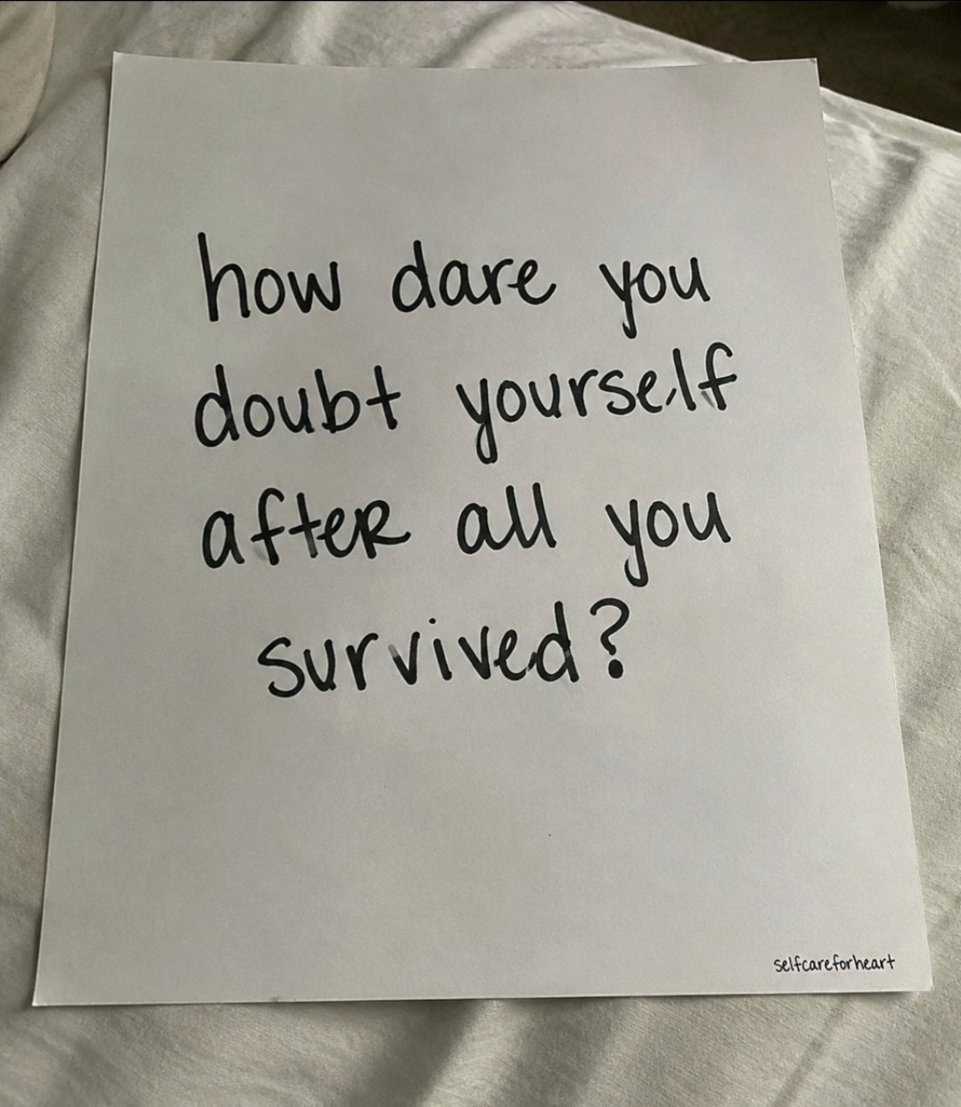 A vertical shot of a white piece of paper resting on a white, slightly wrinkled fabric surface. The paper features handwritten-style black text in the center that reads 'how dare you doubt yourself after all you survived?'. In the bottom right corner, there is a small watermark that says 'Selfcareforheart'.